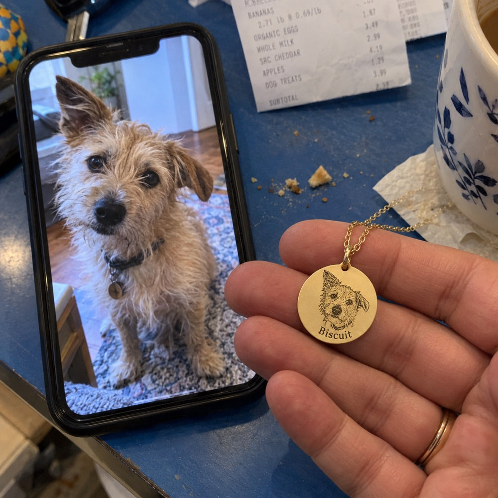 Dog — AI concept: AI demo phone snapshot of a hand holding a gold Biscuit necklace beside a matching terrier reference photo on a phone, on a cluttered blue kitchen table. Not a customer photograph.