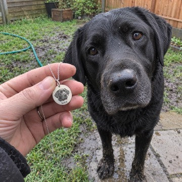AI concept: A hand holds a silver necklace engraved MAX and a matching black Labrador portrait beside the dog in a muddy garden.