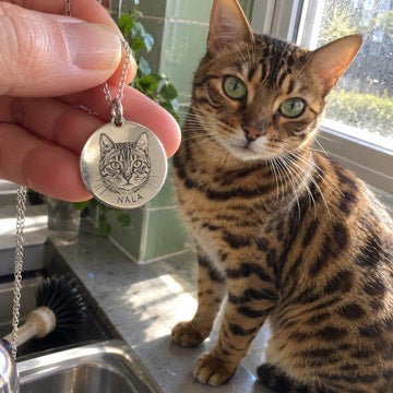 AI concept: Nala the Bengal at a green-tiled kitchen window beside a matching silver engraved portrait pendant.